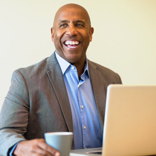 A man in a suit smiles while sitting at a desk with a laptop.