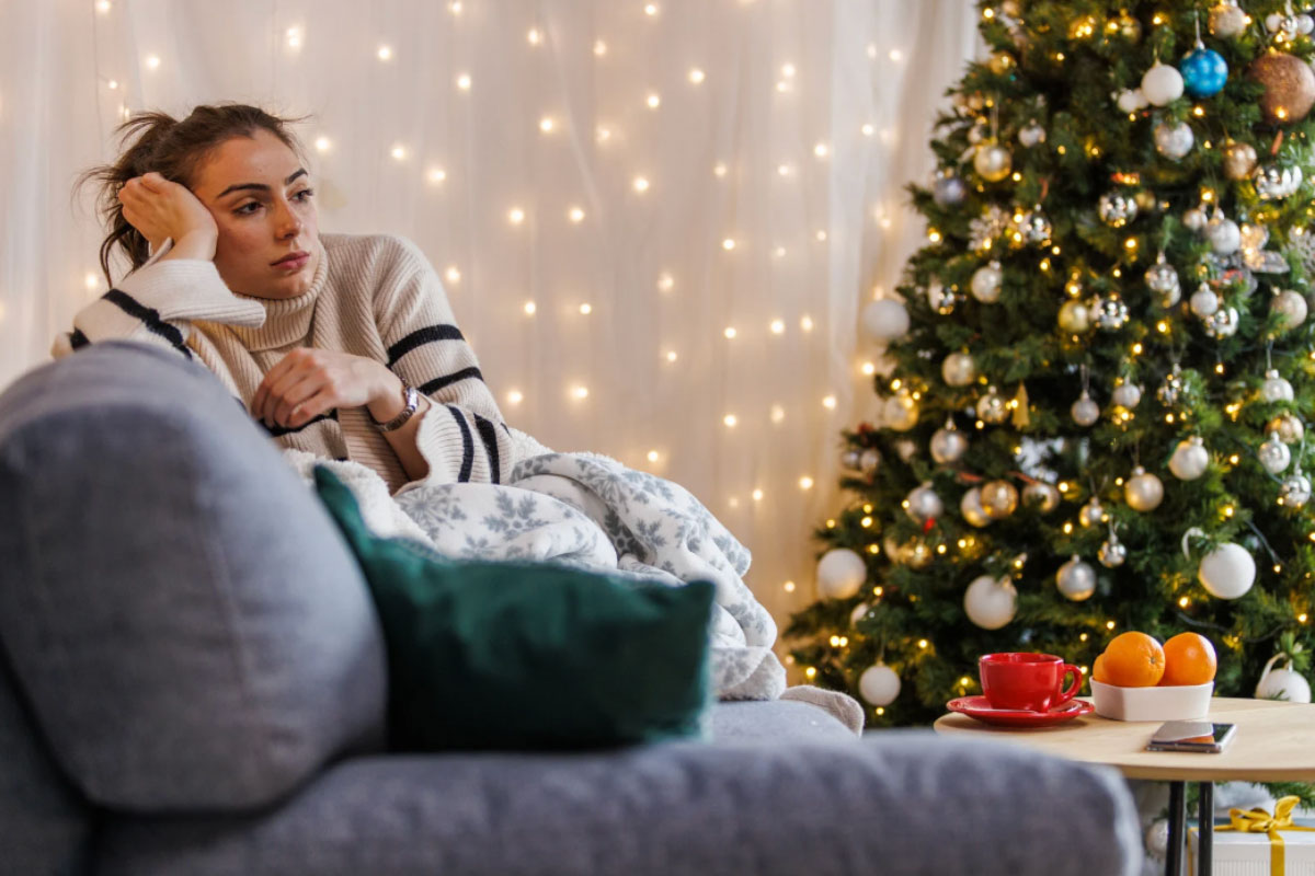 A woman sits on a couch, smiling in front of a beautifully decorated Christmas tree, creating a festive atmosphere.