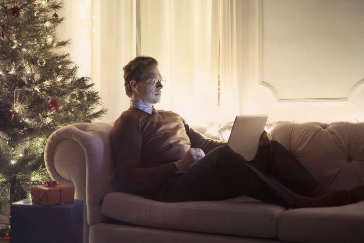 A man sits at a laptop, focused on his work, with a beautifully decorated Christmas tree in the background.