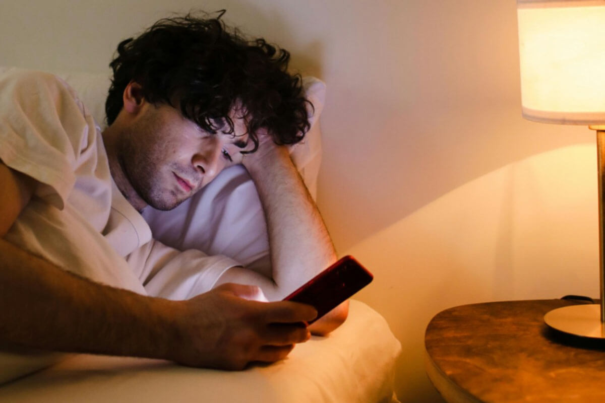 A man lying in bed under warm lamp light, holding a smartphone and looking at it with a contemplative expression.