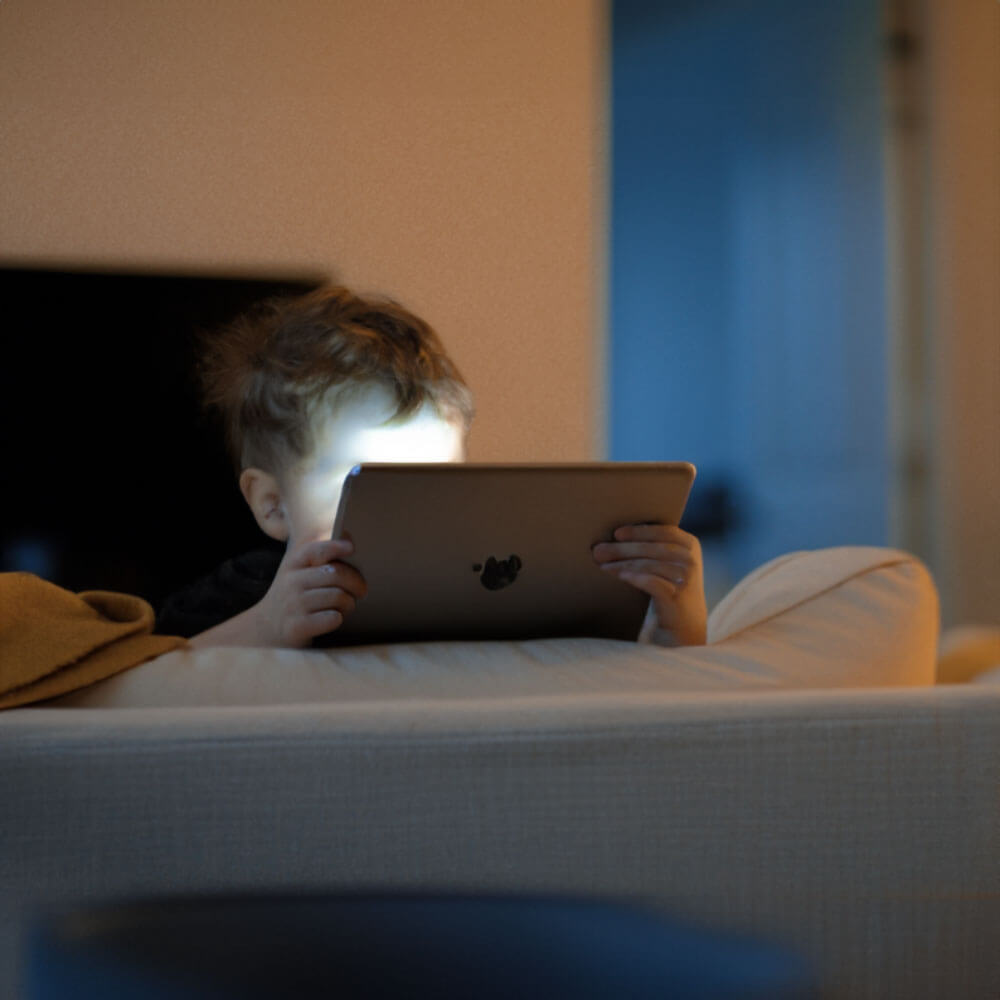 A young child sitting on a couch in a dimly lit room, holding a tablet with its screen glowing brightly against their face.