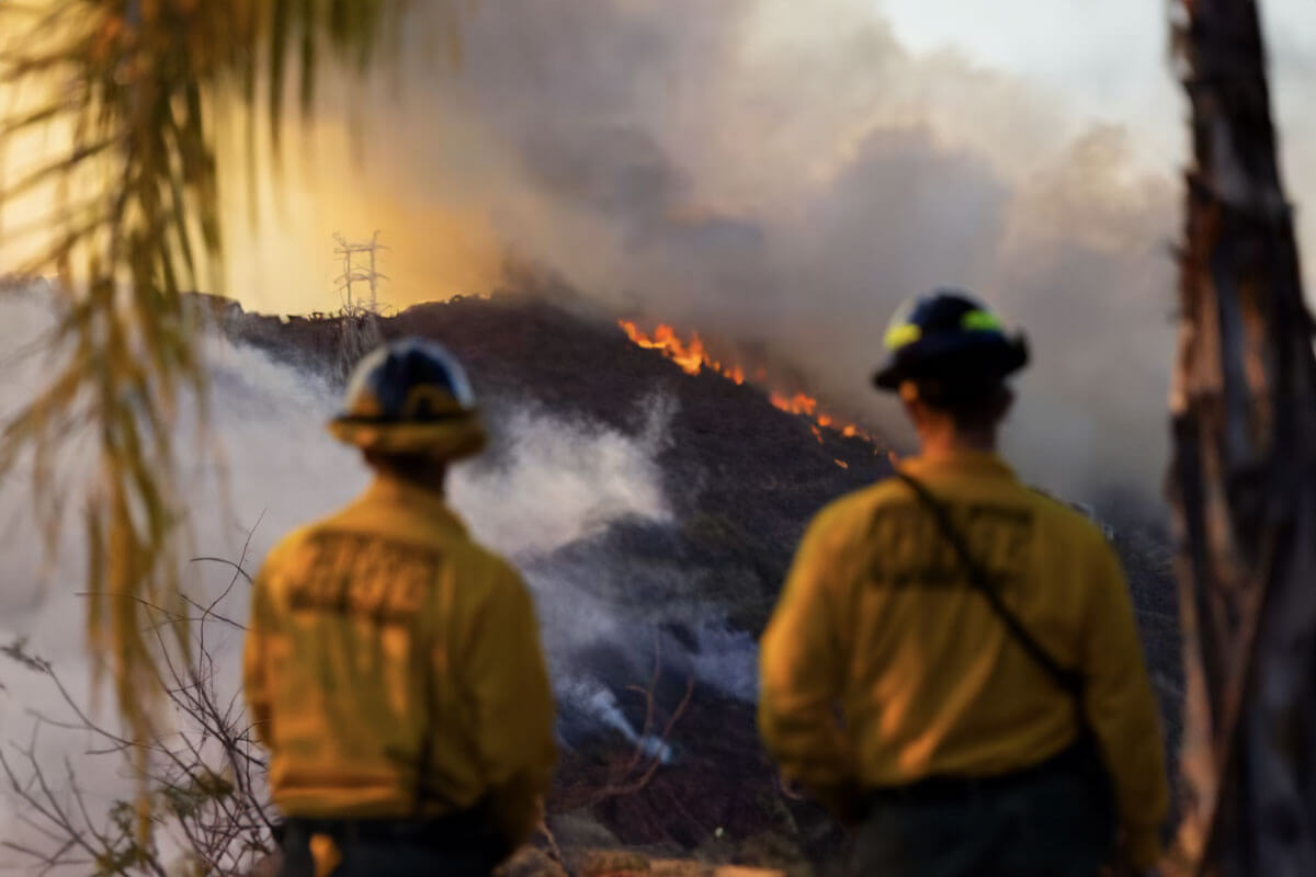 Two firefighters in uniform observing a wildfire on a smoky hillside, with flames visible in the background.