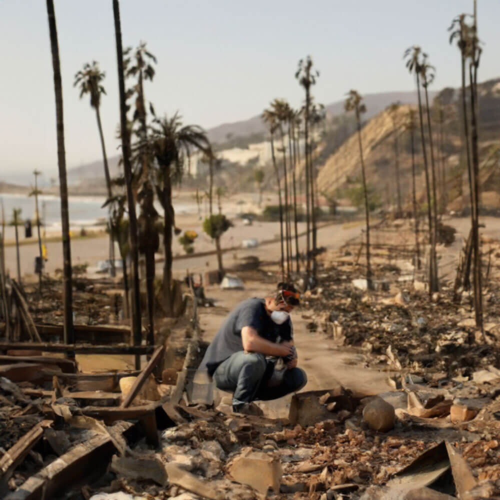 A person kneeling among the charred remains of a fire-ravaged landscape, surrounded by burned structures and scorched palm trees.