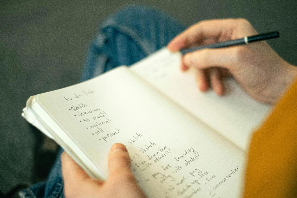 A person writing a to-do list in a notebook while holding a black pen, sitting with crossed legs.