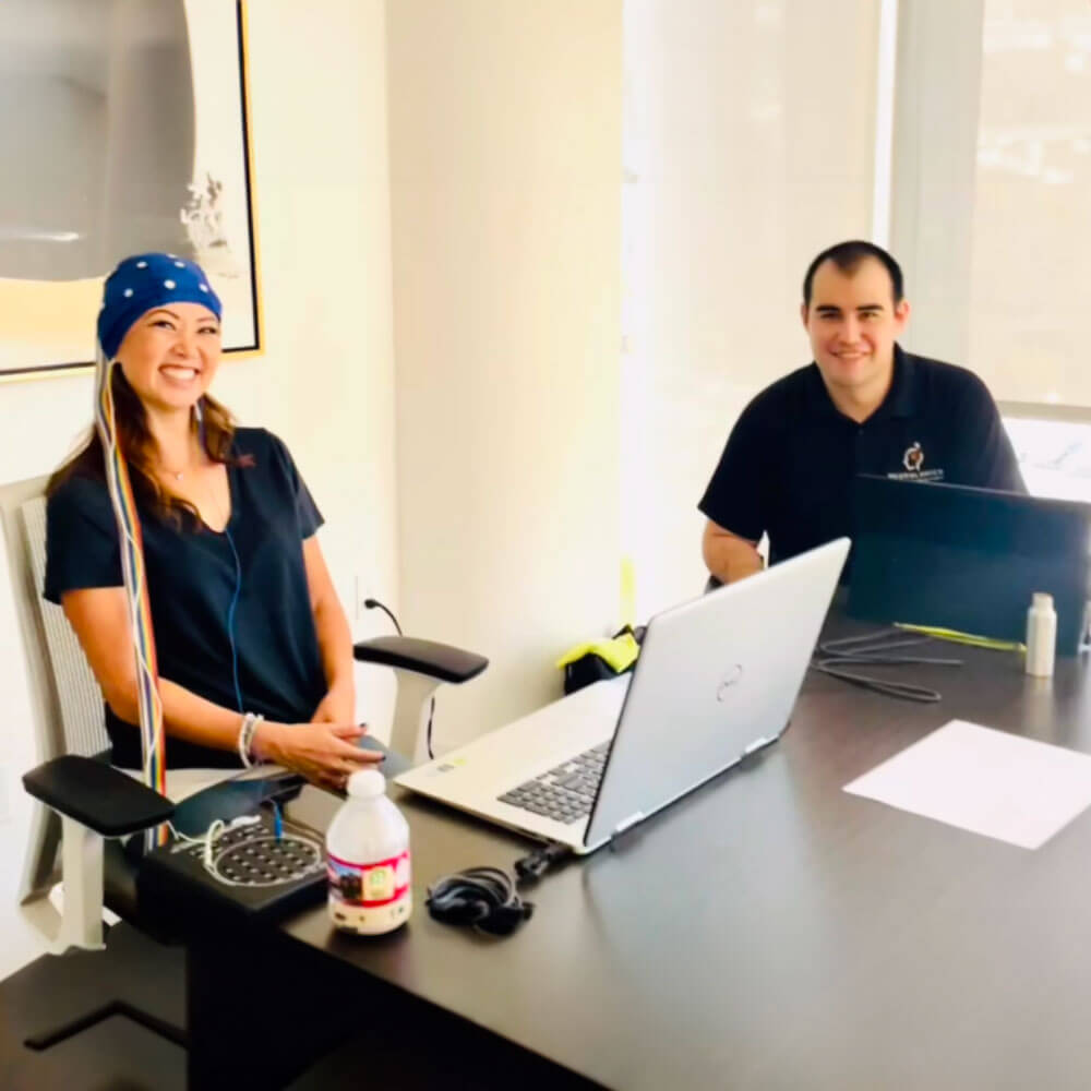 A smiling woman wearing an EEG cap with electrodes, sitting at a desk next to a male colleague in an office setting.