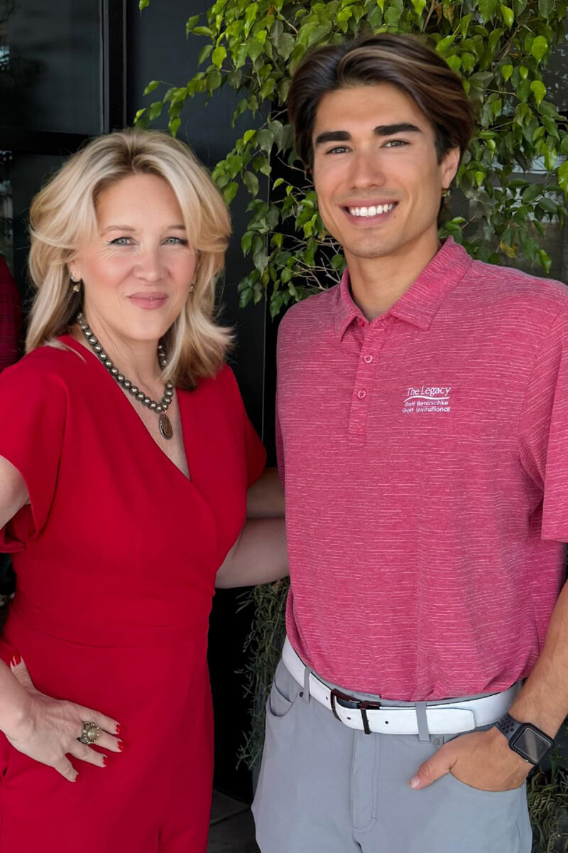 A friendly portrait of two people smiling outdoors, dressed in coordinated red attire. The background includes greenery, giving the image a welcoming and personal tone.