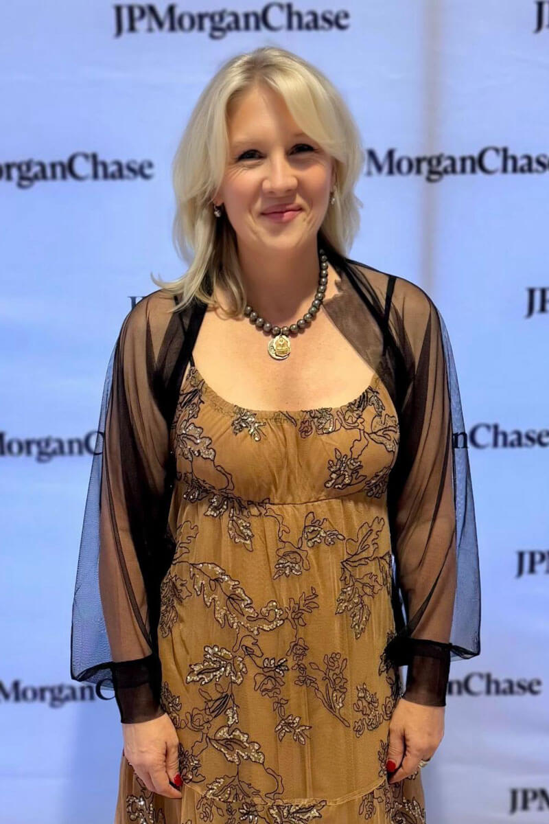 Smiling woman in a tan floral dress with a sheer black shawl standing in front of a JP Morgan Chase backdrop.