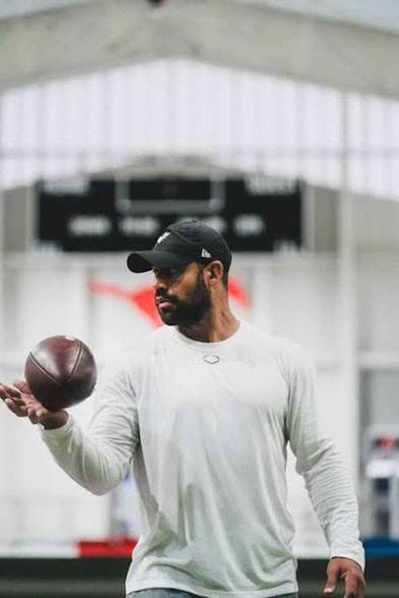 Man in a white long-sleeve shirt and black cap casually tossing a football inside an indoor training facility.