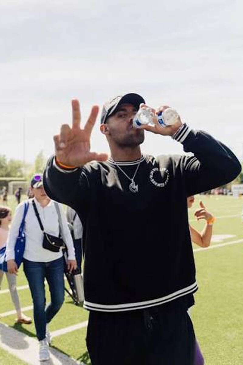 Man wearing a black sweatshirt and cap drinking water while gesturing toward the camera on an outdoor athletic field.