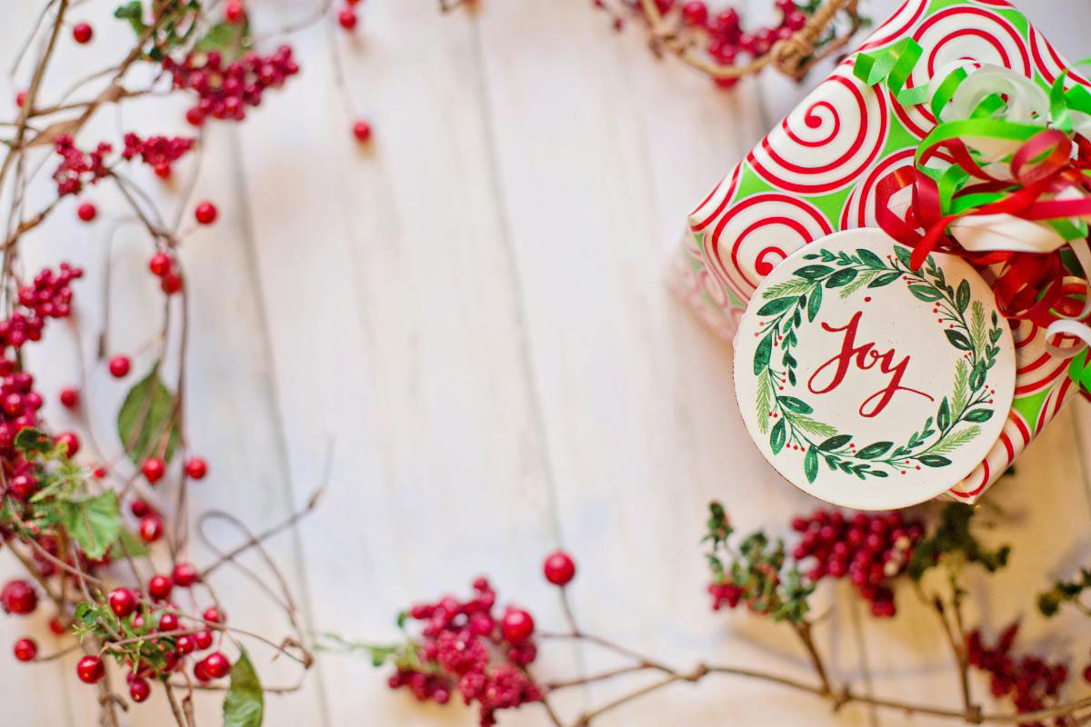 A festively wrapped holiday gift with a “Joy” tag sits surrounded by red berries on a light wooden surface.