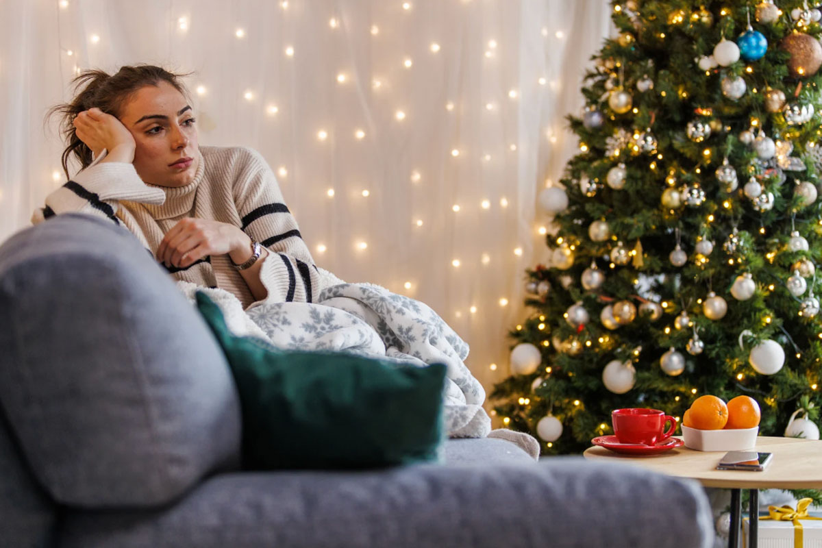 A woman sits on a couch looking sad or reflective beside a decorated Christmas tree and warm holiday lights.