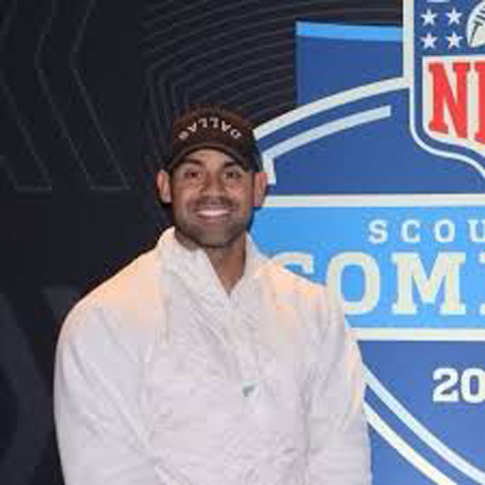 A man in a white jacket and black cap smiles in front of an NFL Scouting Combine backdrop.