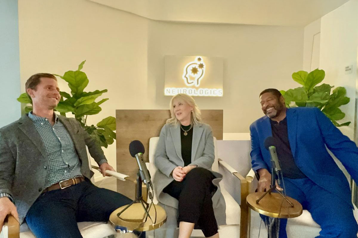 Three people sit together laughing in a modern studio setting with microphones in front of them.