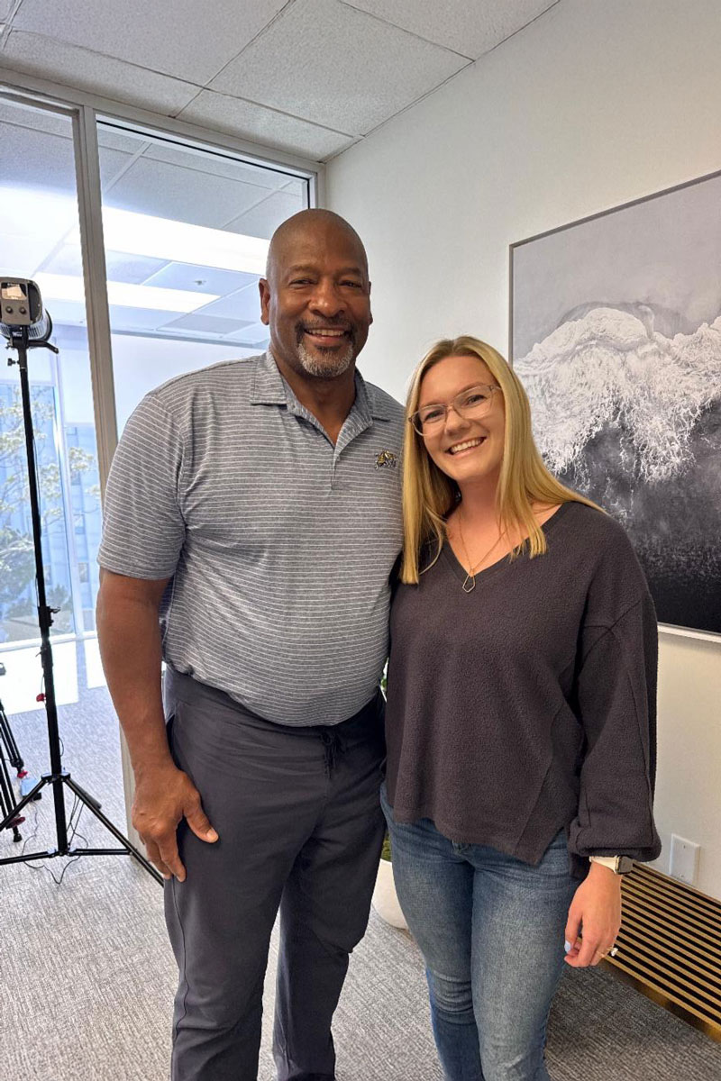 A man and a woman smile together in an office setting with professional lighting equipment behind them.