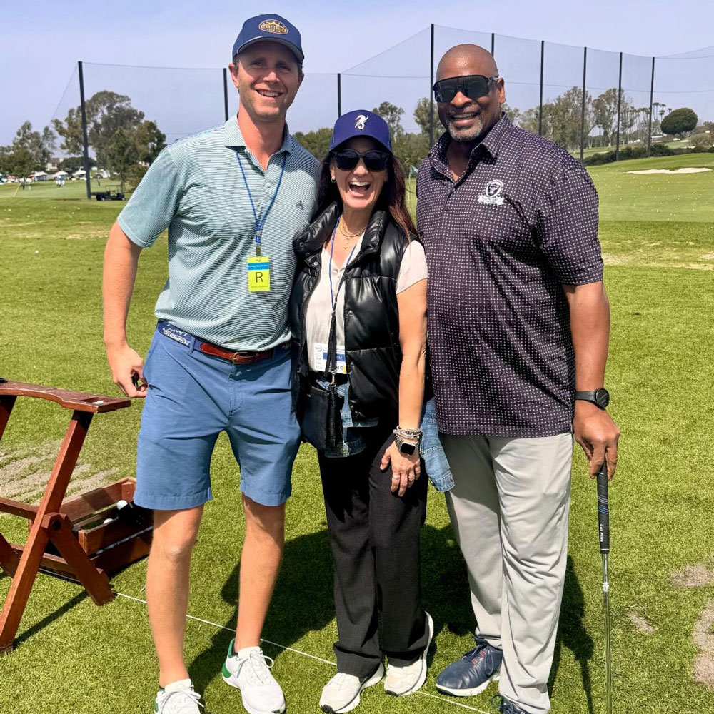 Three people stand together on a golf course smiling for a photo, with one holding a golf club.