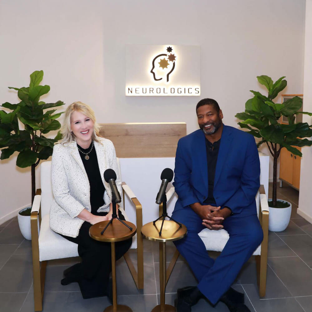Two smiling people — a woman in a white blazer and a man in a blue suit — sit in armchairs with podcast microphones in front of the illuminated Neurologics logo in a modern clinic reception area.