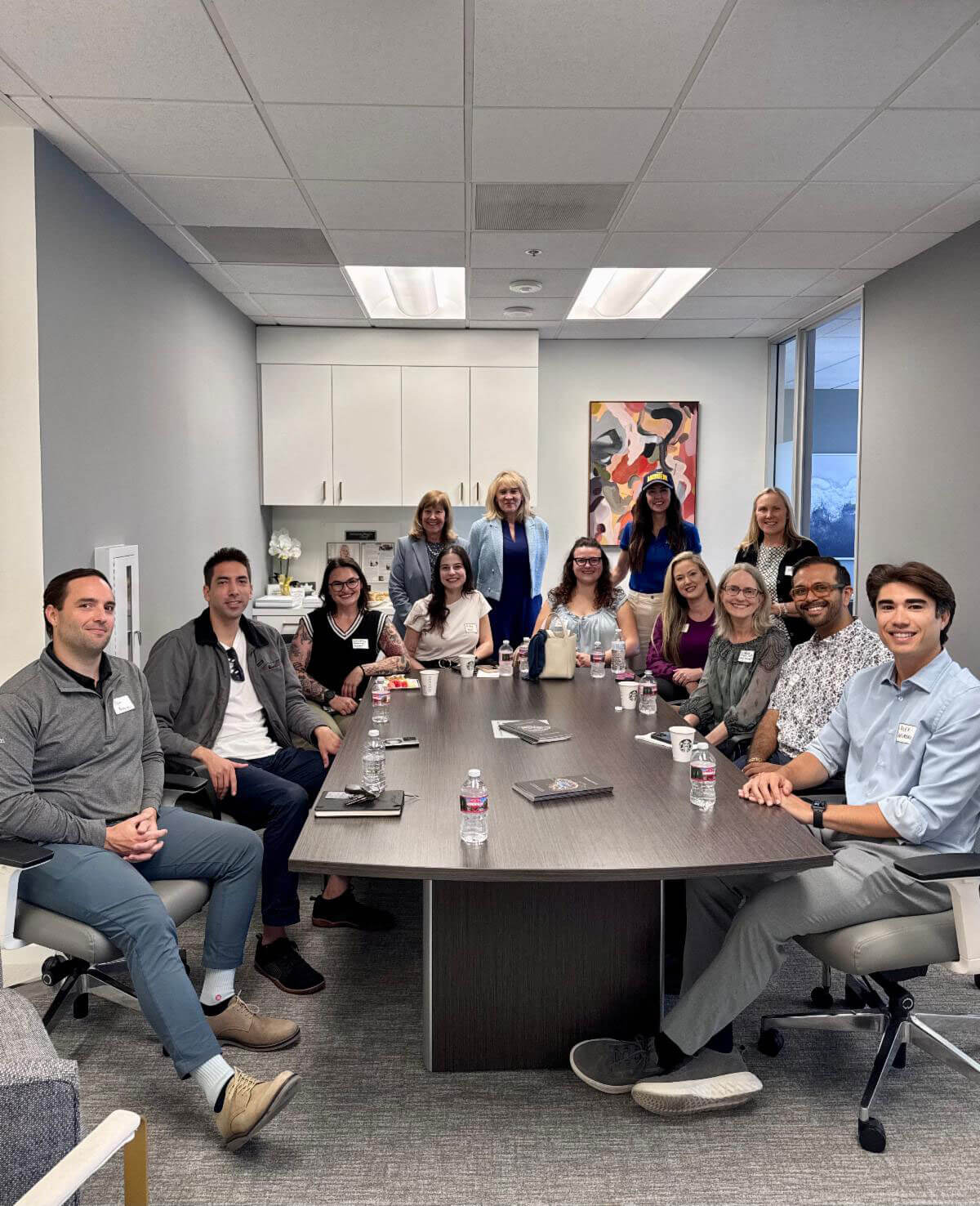 A group of approximately fourteen smiling professionals wearing name tags gather around a large conference table with water bottles and notebooks in a bright modern office.