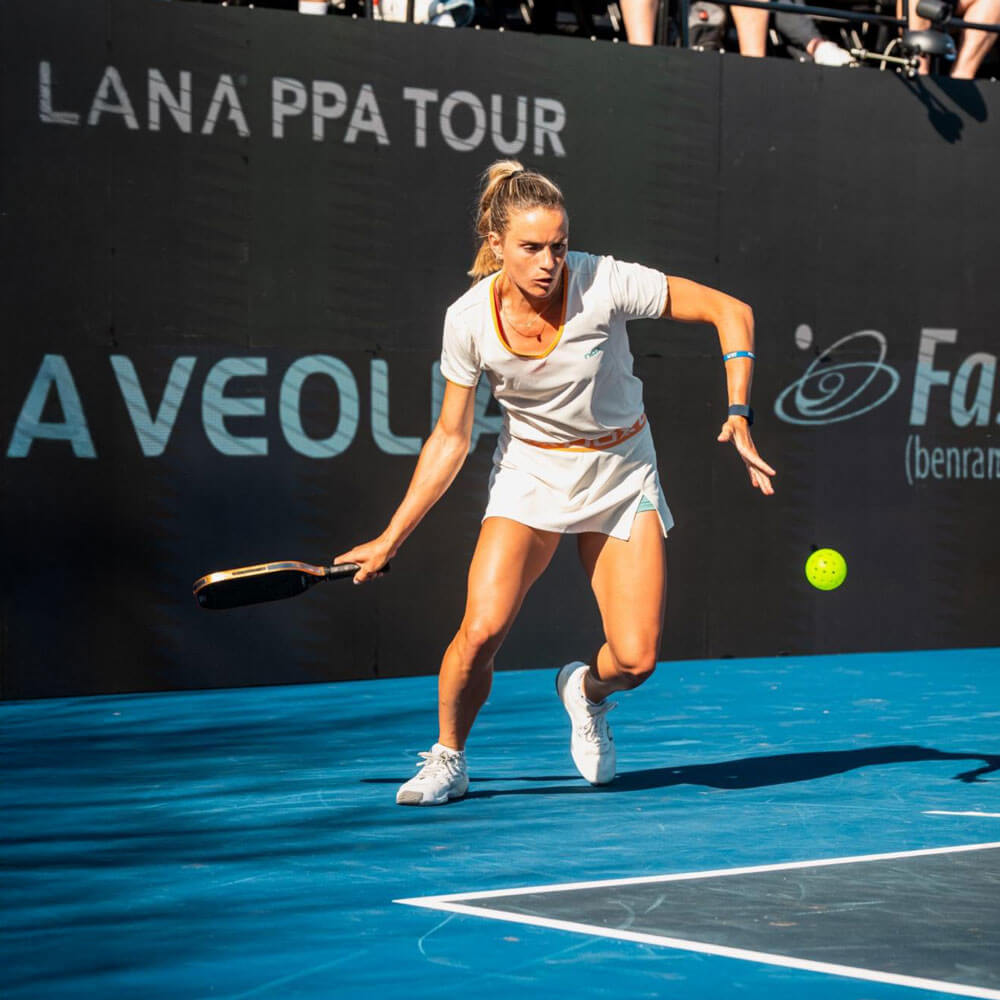 A female pickleball player in white athletic wear lunges forward to return a shot during a professional PPA Tour match on a blue outdoor court.