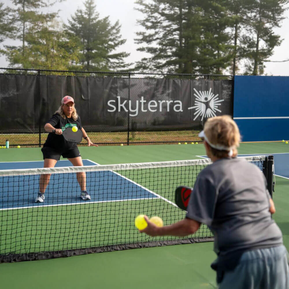 Two women practice pickleball across a net on an outdoor court with a Skyterra-branded fence banner and tall trees in the misty background.