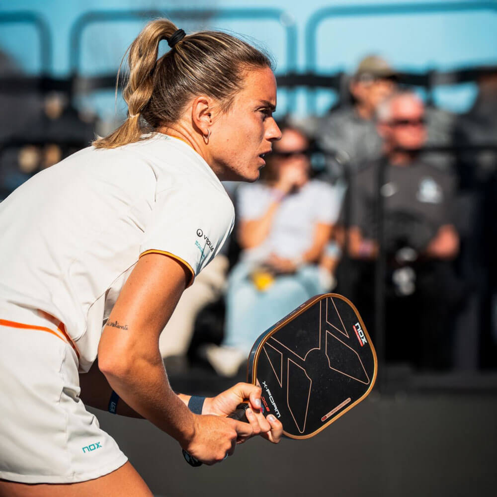 A focused female pickleball player in white and orange athletic wear holds a Nox paddle in a ready stance, photographed in close profile with a blurred spectator crowd behind her.