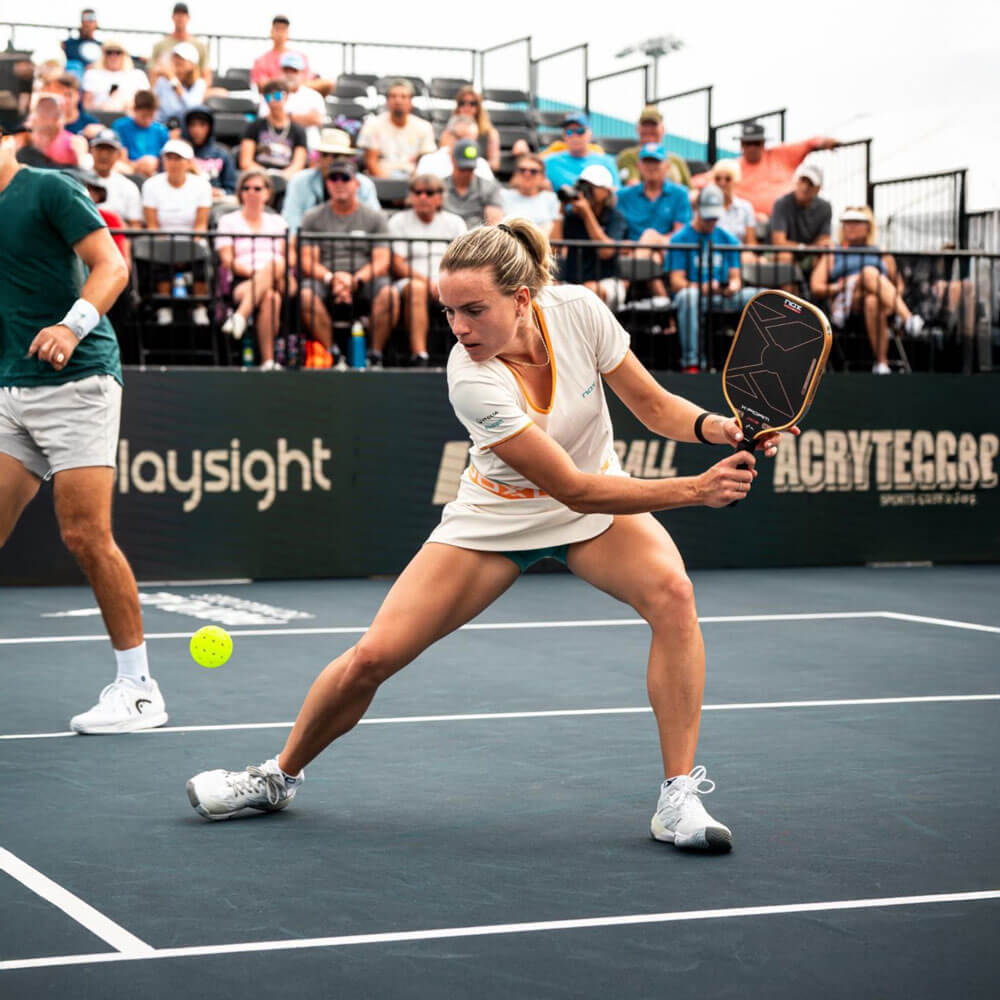 A female pickleball player in white stretches low to reach a ball during a professional doubles match on a dark court, with a large seated crowd visible in the stands.