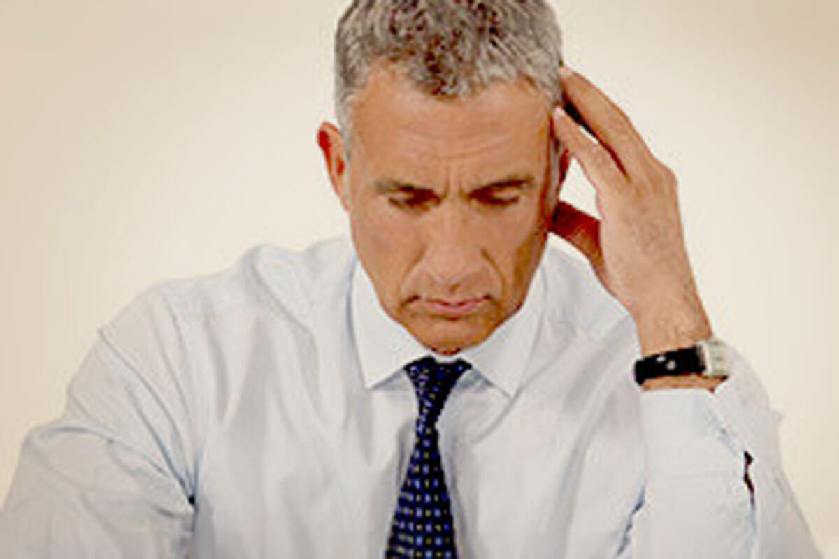 june_newsletter_2026_3 A middle-aged man in a shirt and tie looks down with a furrowed brow, pressing his hand to his temple as if experiencing stress or a headache.