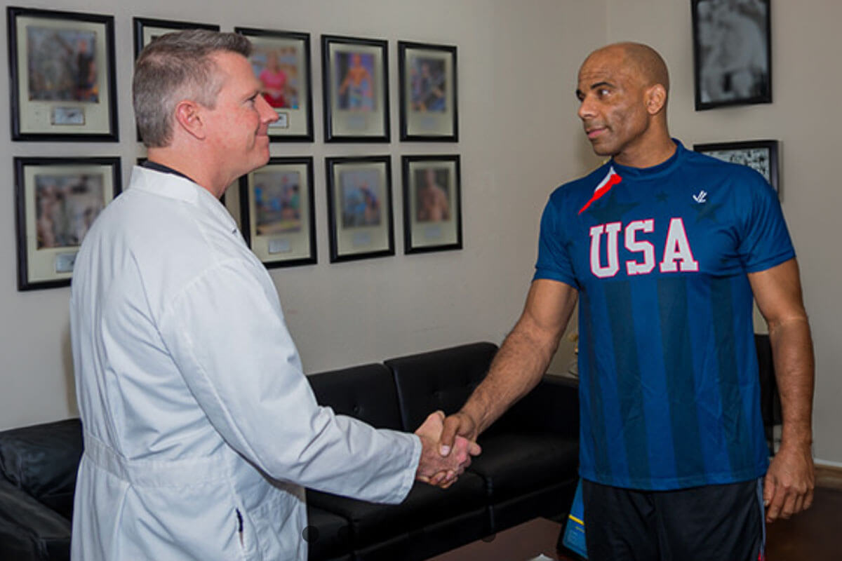 june_newsletter_2026_4 A man in a white coat shakes hands with an athlete wearing a USA jersey inside an office with framed sports photos on the wall.