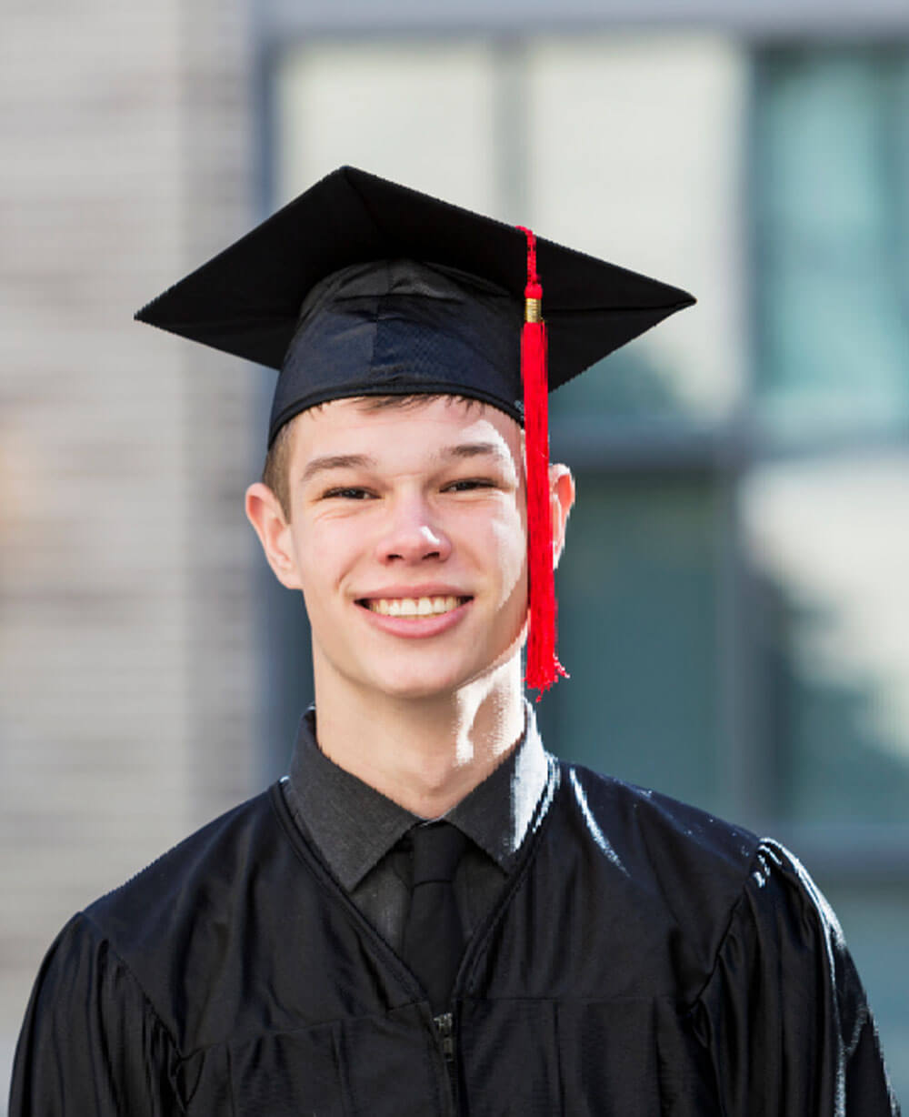 june_newsletter_2026_6 A smiling young man in a black graduation gown and cap with a red tassel poses outdoors in bright sunlight.