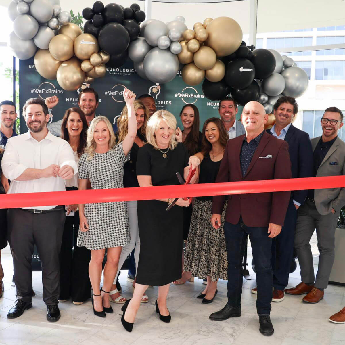 Group of people cutting a red ribbon under a black gold and silver balloon arch at a Neurologics event.