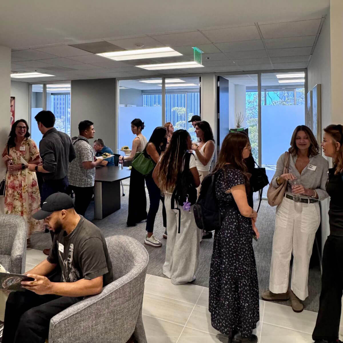 july_newsletter_2026_10 Several small groups of people stand around chatting and networking in a brightly lit office corridor during an event.