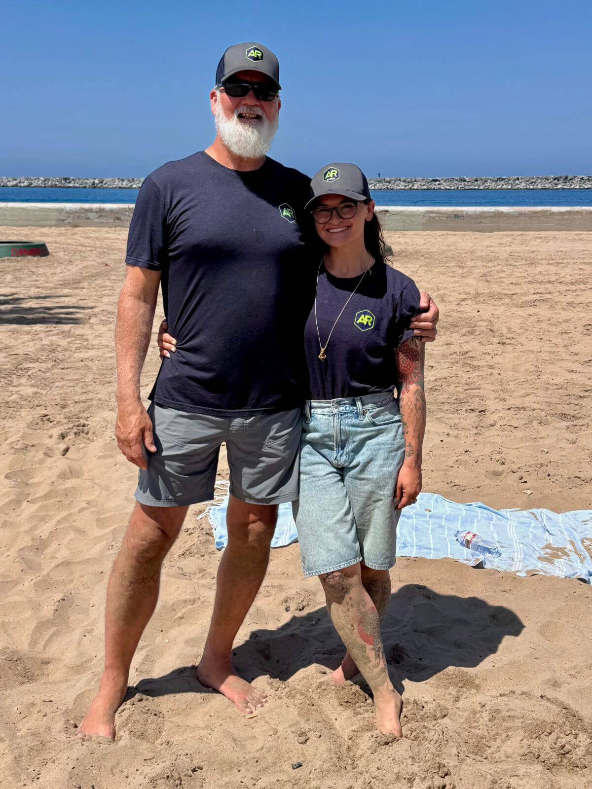 july_newsletter_2026_11 A man with a white beard and a smiling woman stand together on a sandy beach wearing matching dark t-shirts and gray baseball caps featuring an "AR" logo.