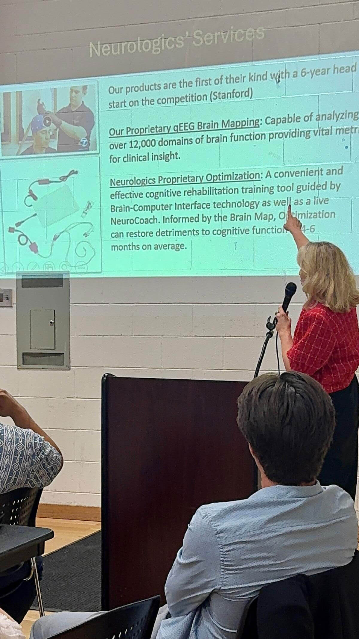 july_newsletter_2026_12 A woman holding a microphone points to a projected presentation slide detailing Neurologics' proprietary qEEG Brain Mapping and Optimization services while addressing an audience.