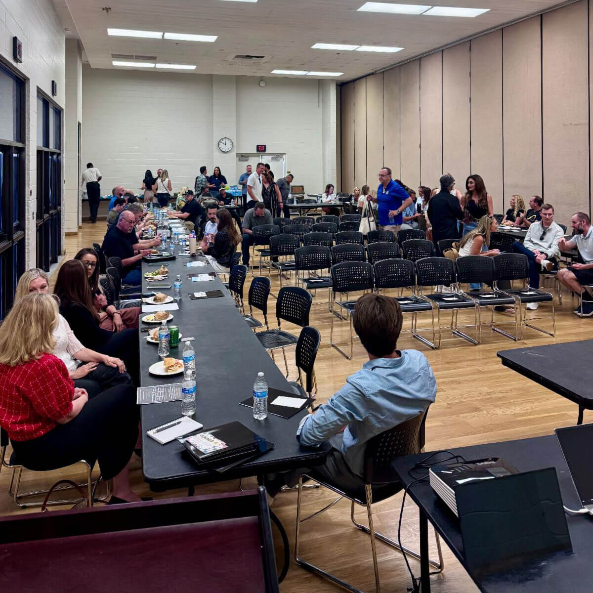 july_newsletter_2026_13 Dozens of attendees sit at long tables eating a catered meal and socializing in a spacious, brightly lit hall during a community event.