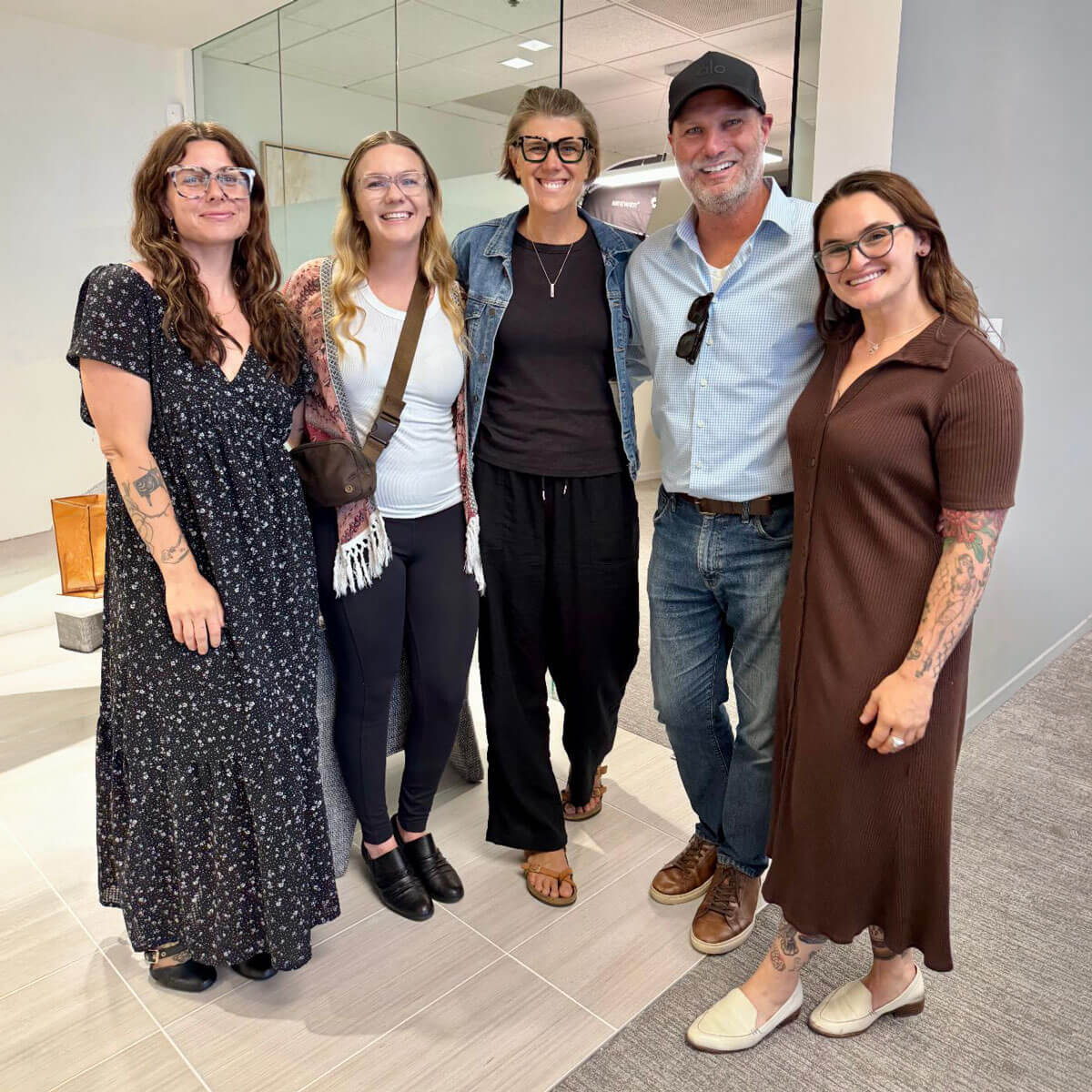 july_newsletter_2026_4 Five casually dressed professionals standing together and smiling warmly for a photo in a modern office hallway.