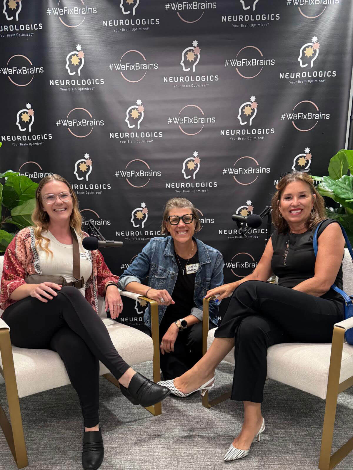 july_newsletter_2026_7 Three smiling women sit comfortably in armchairs around a microphone in front of a Neurologics step-and-repeat banner.
