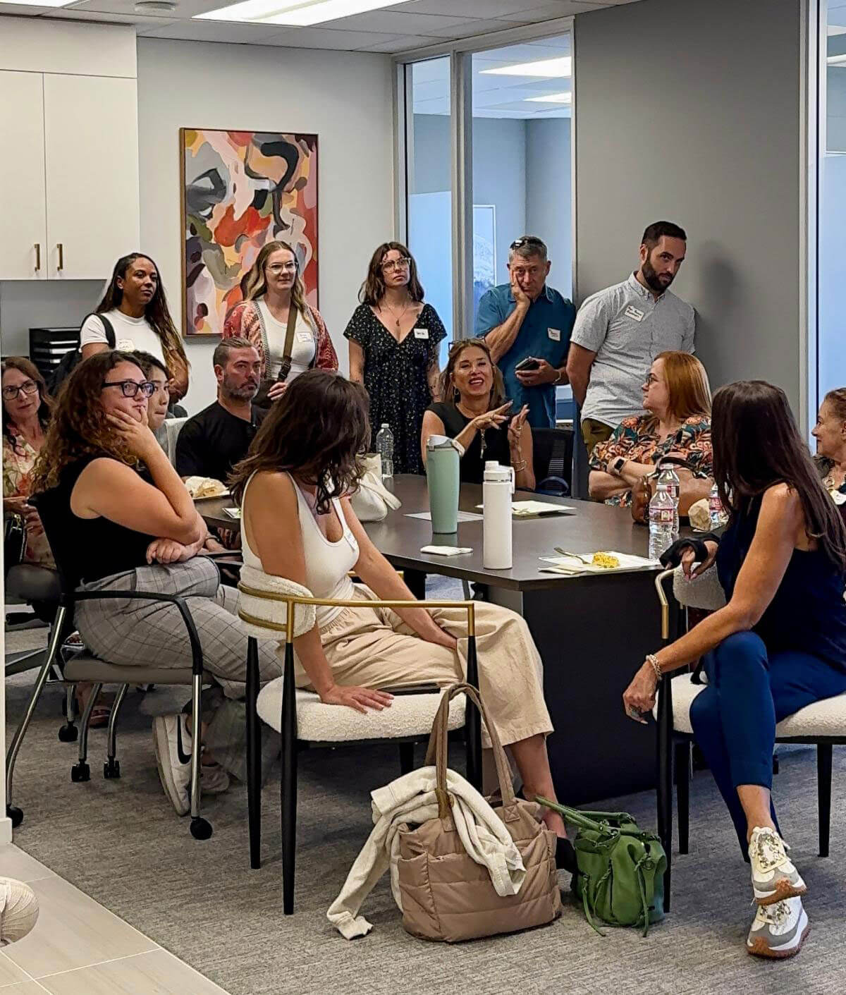 july_newsletter_2026_8 A diverse group of event attendees sit and stand around a conference room table, listening attentively to an off-camera speaker.