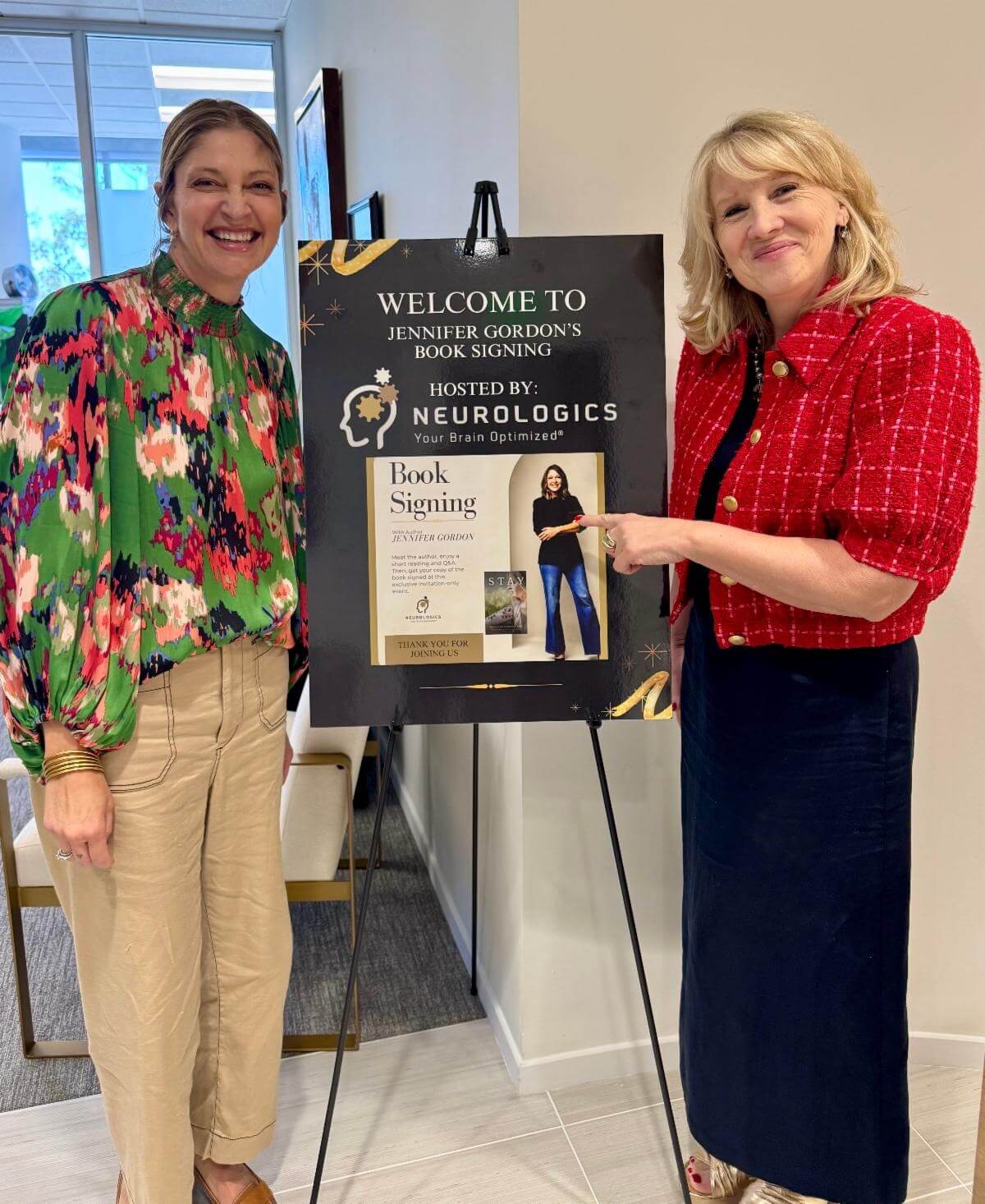 Two smiling women stand beside an easel sign reading "Welcome to Jennifer Gordon's Book Signing" hosted by Neurologics.