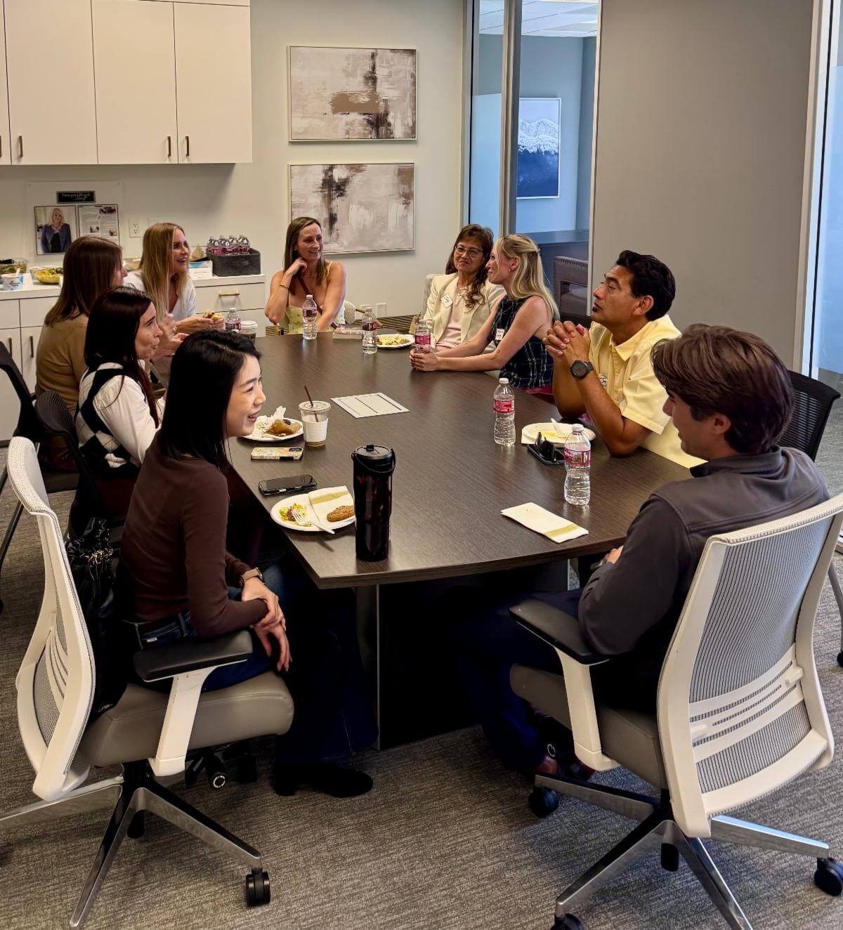 A group of people sit around a conference table with plates of food, chatting in a bright office meeting room.