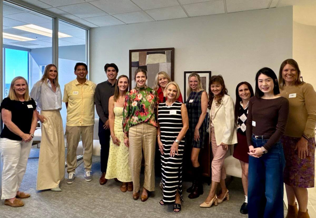 A group of thirteen smiling people in business casual clothing pose together in an office with large windows.