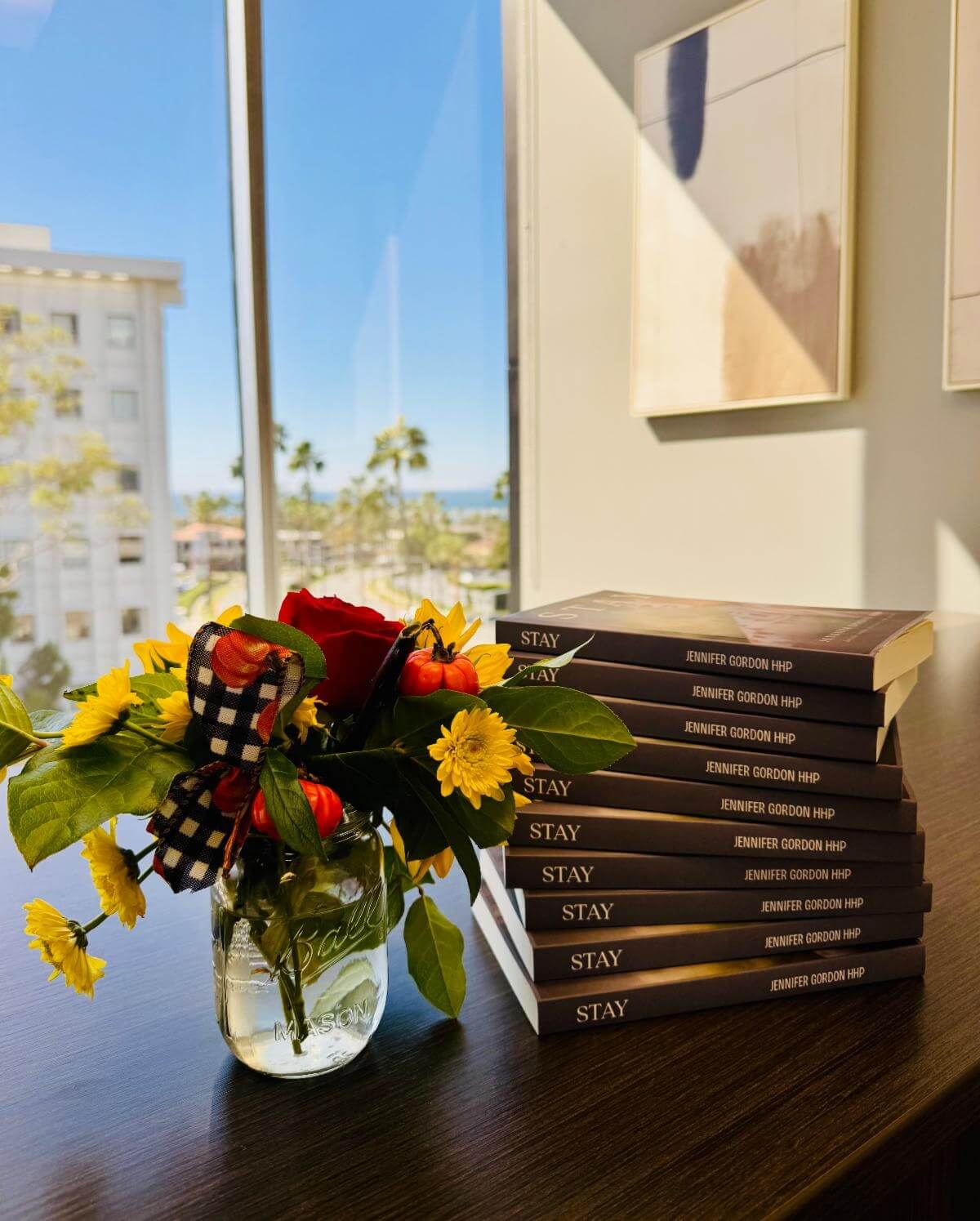 A stack of paperback copies of "Stay" by Jennifer Gordon HHP sits beside a mason jar of yellow flowers and small pumpkins.