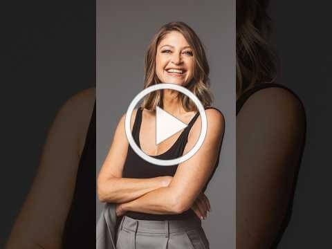A smiling woman in a black tank top and gray trousers stands with her arms crossed, with a video play button over the image.