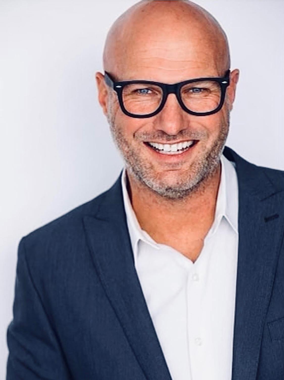 A smiling bald man with black rimmed glasses and a gray beard wears a navy blazer over a white open collar shirt.